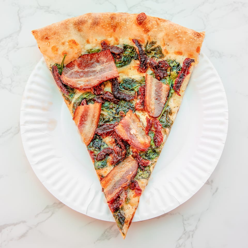 Triangular flatbread topped with bacon, spinach, and sun-dried tomatoes on a white paper plate.