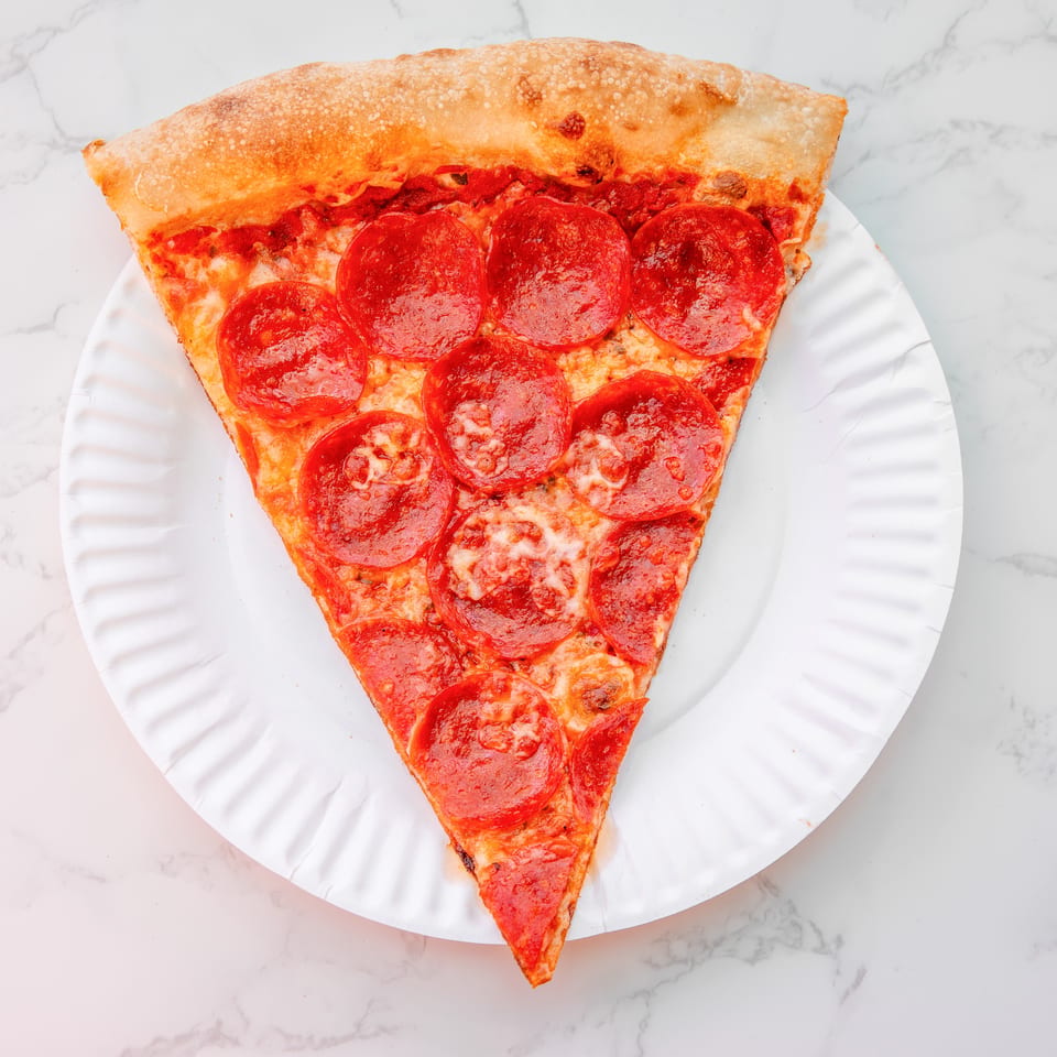 Large triangular pepperoni pizza slice on a white paper plate over a marble surface.