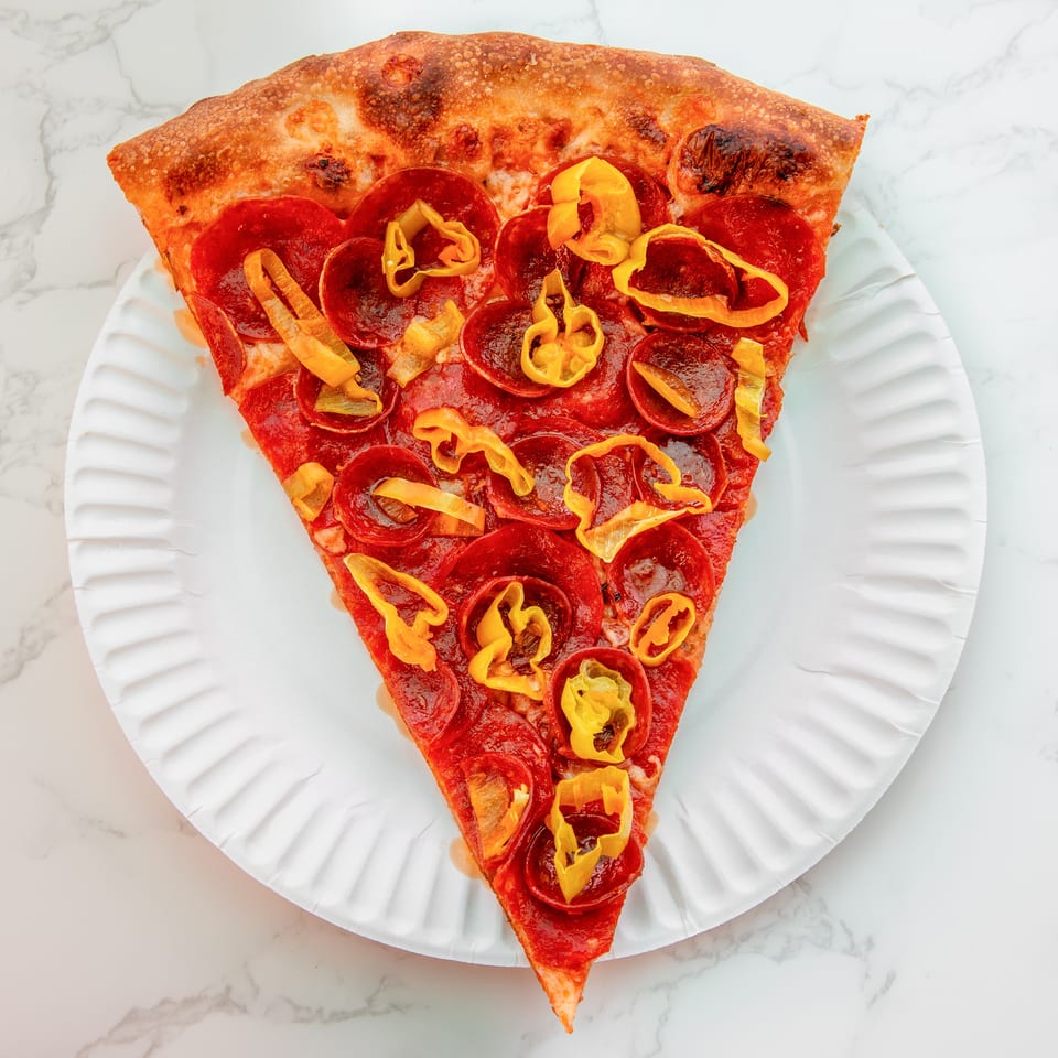 Triangular pepperoni pizza slice topped with yellow peppers on a white plate over a marble surface.