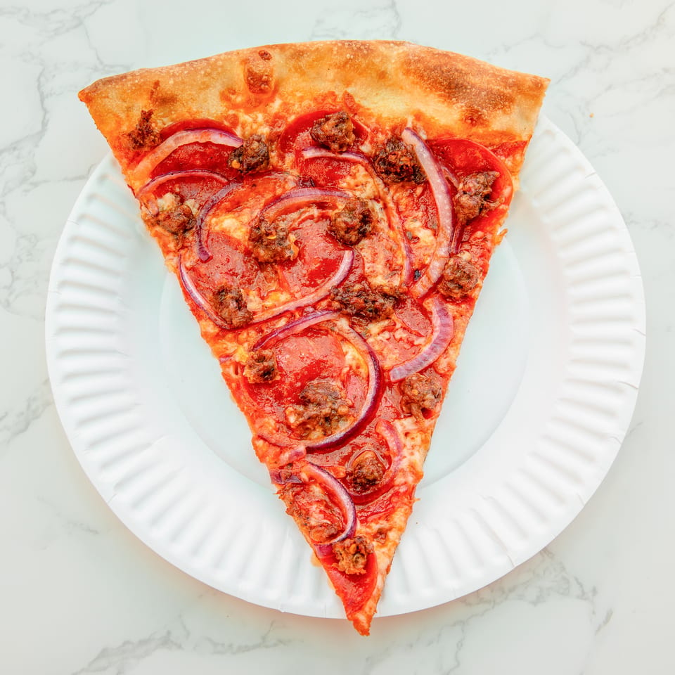 Triangular slice of pepperoni and sausage pizza topped with red onion on a white plate.