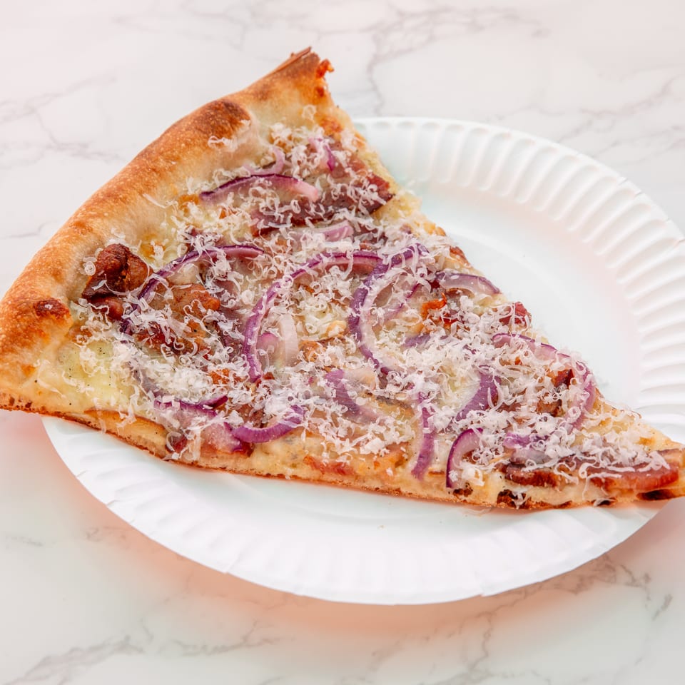 Triangular slice of cheesy pizza topped with red onion, sausage, and grated cheese on a white plate.