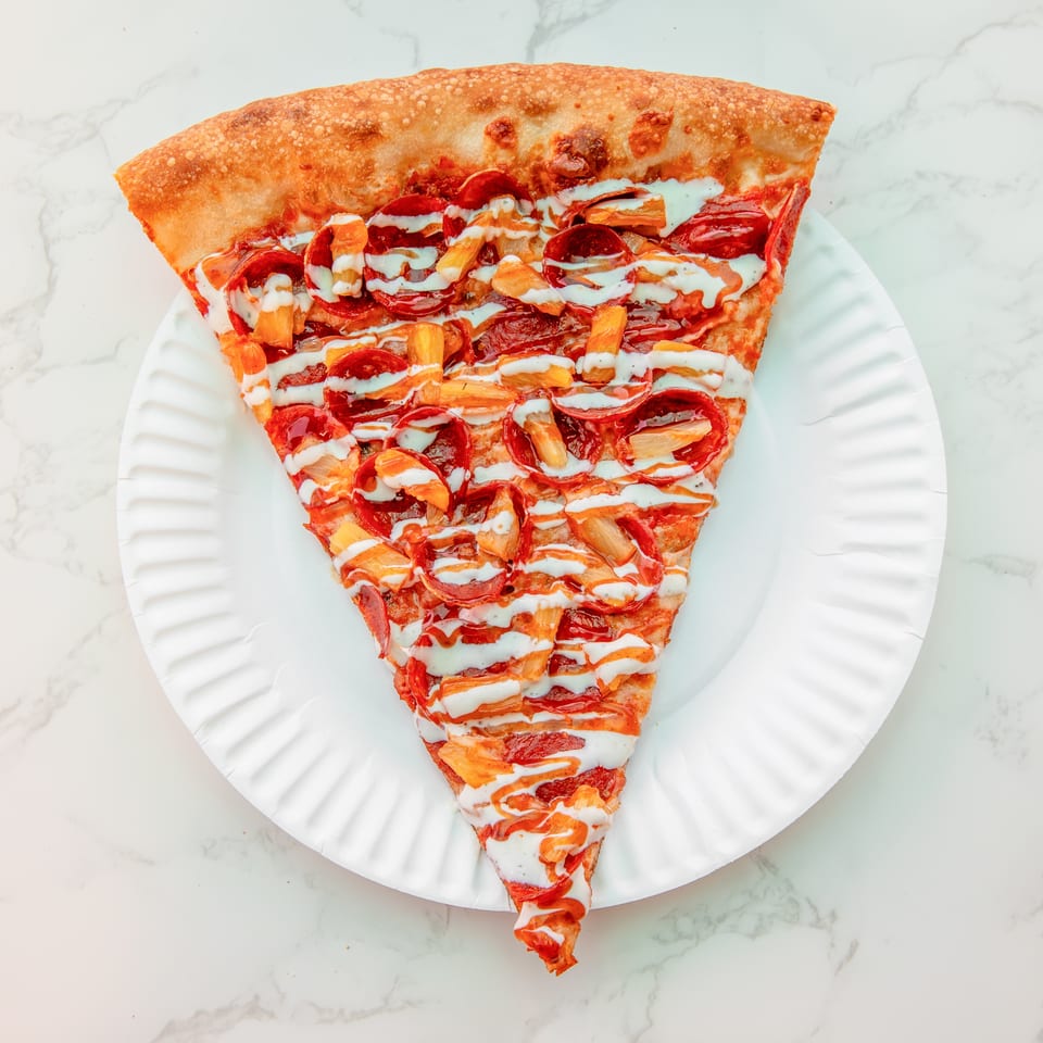 Triangular pepperoni pizza slice topped with pineapple chunks and drizzled white and orange sauces on a white plate.