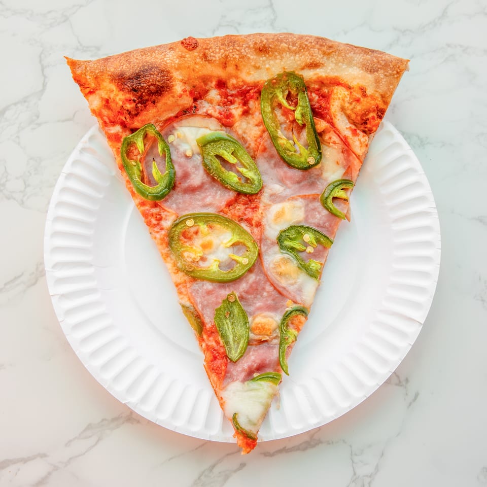 Slice of pepperoni pizza topped with green jalapeño peppers on a white plate against a marble surface.