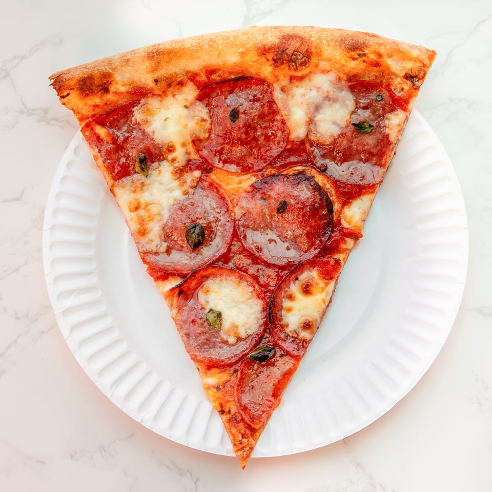 Pepperoni pizza slice with melted cheese and basil on a white plate over a marble surface.