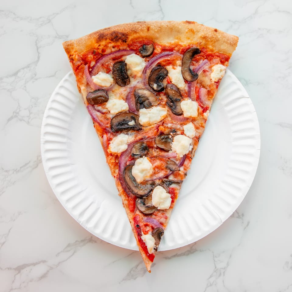Triangular pizza slice topped with mushrooms, red onions, tomato sauce, and white cheese on a white plate.