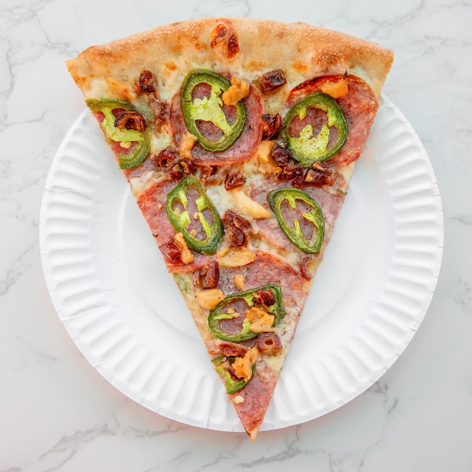 Triangular slice of cheesy pepperoni pizza topped with jalapeños and roasted vegetables on a white plate.