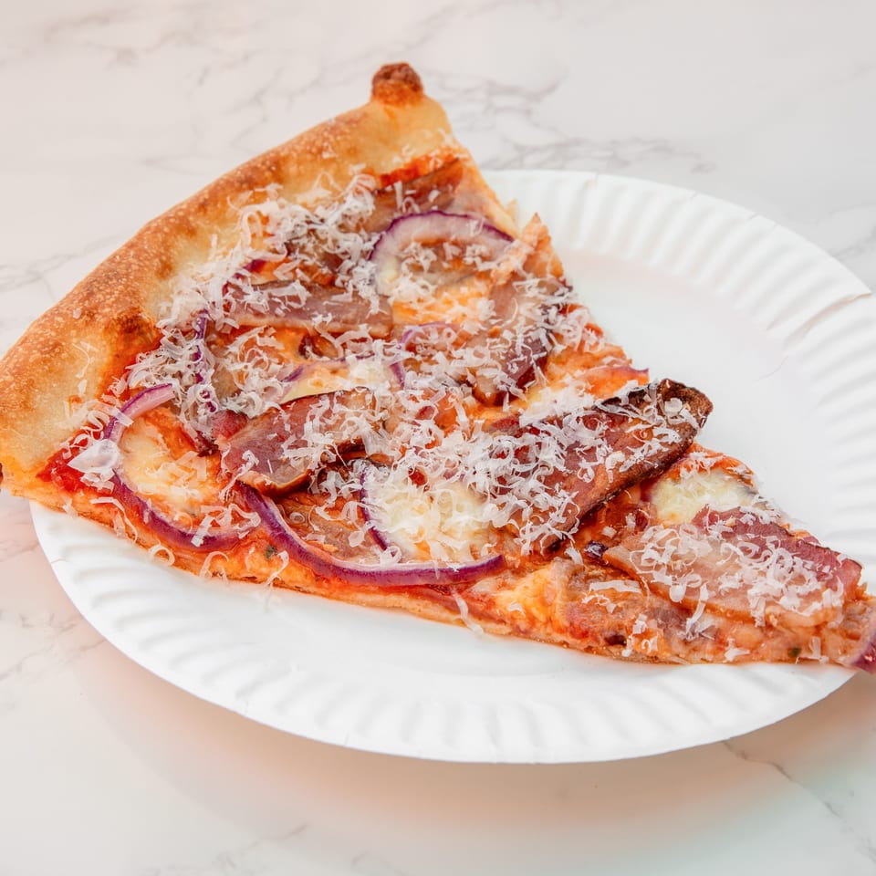 Large triangular pizza slice topped with melted cheese, cured meat, red onion, and grated cheese on a white paper plate.