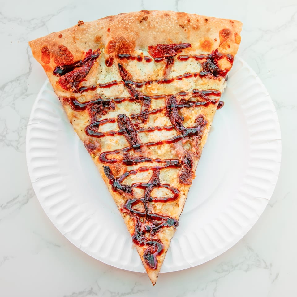 Triangular flatbread topped with melted cheese, sun-dried tomatoes, and balsamic glaze on a white plate.