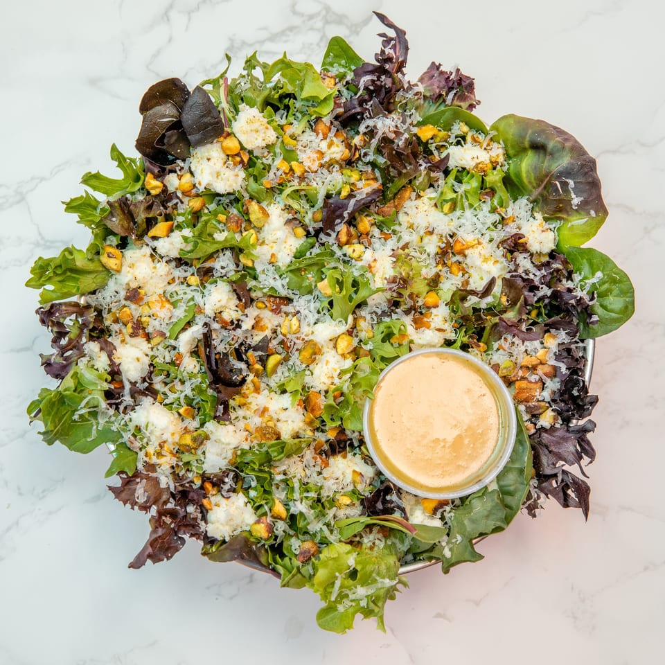 Mixed green salad topped with shaved cheese, pistachios, creamy dressing, and white cheese on a marble surface.